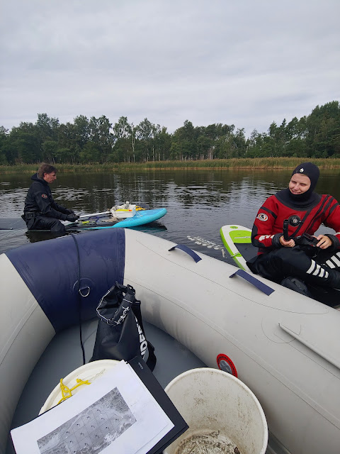 Kaksi henkilöä kuivapuvuissa, kumivene ja SUP-lauta