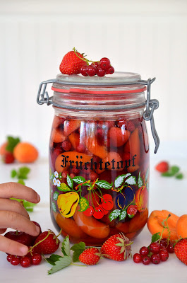Ein großes Einmachglas mit dem Aufdruck Früchtetopf ist mit frischem Obst und Rum gefüllt. Rundherum liegen Früchte dekorativ arrangiert. Eine Kinderhand greift nach einem Stück.