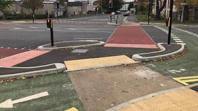 The floating pedestrian island of a CYCLOPS junction. There is a signalised crossing of the road to the left and ahead with a cycle track crossing in the foreground.ead