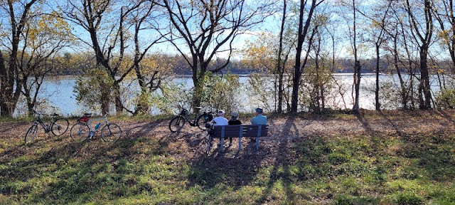 Four people sitting on bench overlooking tree-lined river with bikes parked nearby.