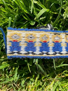 A short section of tablet woven band showing little yellow men in blue overalls, photographed against a grassy background