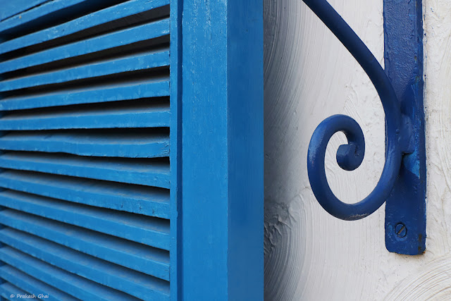 A Minimalist Photograph of a Blue Metal Curl Shown in contrast with Simple Lines of a Window.