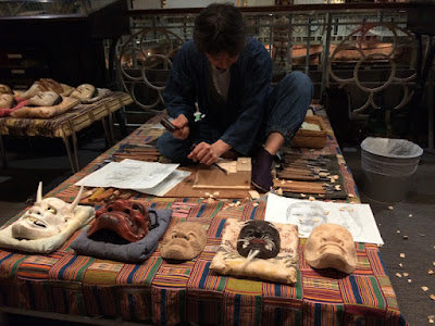 Man sitting cross legged on a  small stage carving a mask.