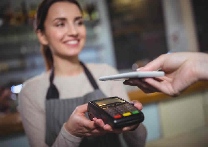 Imagem mostra uma mulher segurando uma maquina de cartão de crédito e outra pessoa faz o pagamento com um smartphone com NFC.