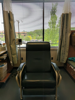 An empty chemo chair with the IV pole in behind it. The window looks out onto spring trees and greenery behind the building.