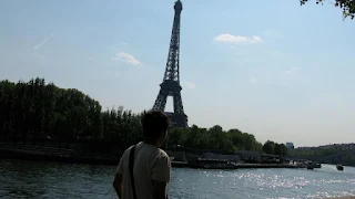Man with his back to the camera. In front of him is a river and on the other side of the river are green trees and the Eiffel Tower