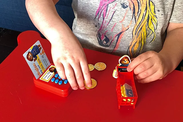 Close up of the toys with the first issue of the paddington bear magazine including a plastic chip and pin machine, mini shopping trolley, foam pieces and a pretend cash card