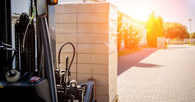 A forklift moving product pallets and boxes out of direct sunlight. The boxes are wrapped in plastic wrap.