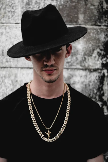 Stylish model in black attire wearing a Fedora hat, golden plated Cuban link chain, and sporting a light beard against a rustic wall backdrop.