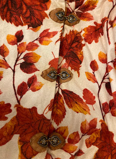 A close-up of three leather-backed buckles on a flannel coat patterned with orange and red autumn leaves on a white background. The buckles are shaped like black and gold spades or hearts, the leather behind them is a pale brown heading towards tan.