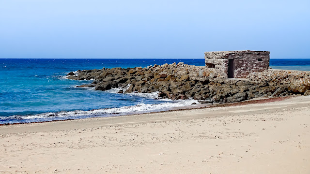 Playa de arena con una escollera con una construcción y las azules aguas del mar.