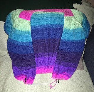 Front view of brightly coloured striped cardigan. The sleeve caps are on hold, as are the bottom body stitches. The stripes are a gradient of pink, mint, teal, blue, and purple shades.