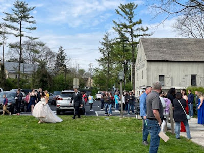 Image shows the parking lot, wooden barn (museum store), and entry walkway at Ephrata Cloister. Adults in casual clothes and teens dressed in formal wear are standing around waiting to take photos.