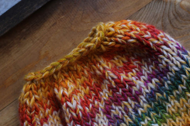 Close-up of the top half of a yellow and rainbow knitted beanie with a hole on the top for a pony tail or bun. It lays on a wooden surface.