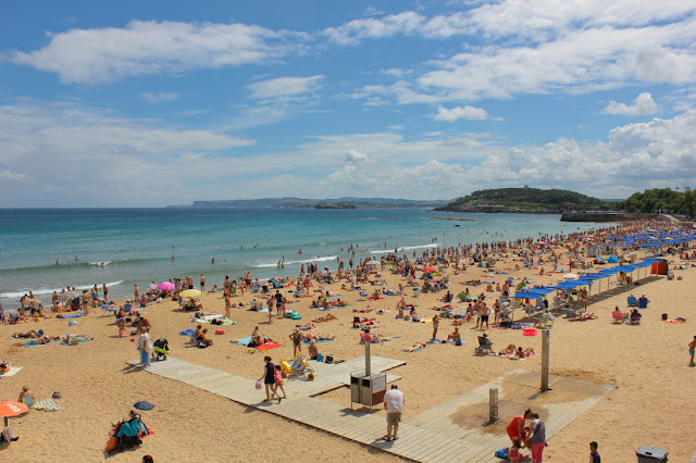 W letnie, lipcowe południe na plaże w dzelnicy Sardinero w Santader. Długa piaszczysta plaża pełna plażowiczów. Wyłożone na piasku ręczniki, ustawione parasole malują się na szarożółtym piasku wieloma kolorowymi punktami. Morze jest spokojne.
