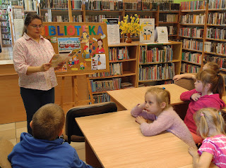 Bibliotekarka stoi na tle regałów z książkami i lady bibliotecznej.  Po lewej stronie  bibliotekarki znajduje się tablica z ilustracjami, obrazującymi zawód bibliotekarza o którym ona opowiada przed nią siedzącej na krzesełkach przy stolikach grupie słuchających ją dzieci z  Zespołu Przedszkolno-Szkolnego nr 3 w Szczakowej.