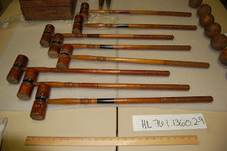 Eight wooden croquet mallets lined up on a table with yardstick - the handles are approximately on yard long, the heads of the mallets have grooves and painted stripes