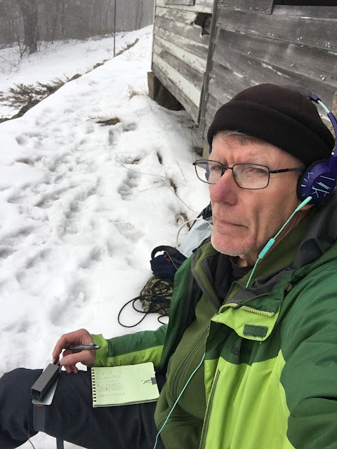 A man in winter sits outdoors leaning on a clapboard-sided shack.