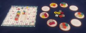 On the left is the scoring board. A track weaves around the outer edge of the board, with four coloured cubes on it to indicate the score of each player. In the centre of the board is a row of the stained glass tiles, and next to that is another track showing negative point values, with four more cubes to track player's broken glass penalties. On the right is a ring of nine discs, some of which have four of the glass tokens on them, and some of which are empty. These discs are arranged in a circle, and in the centre of the circle are some more of the stained glass tokens, along with the first player token.