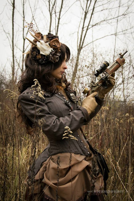 Woman in steampunk clothing in shades of brown, carrying a steampunk gun, wearing a fascinator with a animal skull