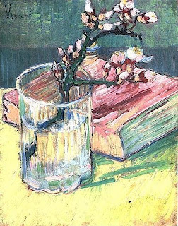  quadro de ramo de amendoa em flor com tom lilás em um copo junto a um livro de capa rosa em cima de uma mesa  