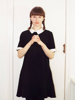 Ellie, with hands clasped looking straight at the camera, wearing a black dress with white collar and cuffs, with hair in plaits.