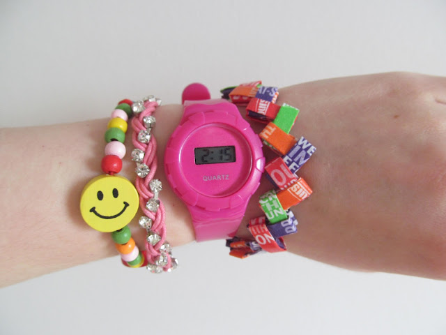 wooden bracelet with multicoloured beads and large yellow smile bead, woven pink rope bracelet with silver gems, hot pink plastic watch and oragami multicoloured zigzag bracelet all on white wrist