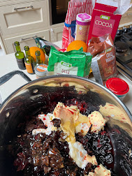 A large pan is centre of the picture. It's filled with ingredients making a cake mixture. There's a lump of butter, a range of dried fruit, cocoa powder, all in a big brown, glossy mulch. Around the pan, are some of the ingredients. A box of cocoa, baking powder, two oranges, a box of eggs, a pack of dates, and some little bottles of port.