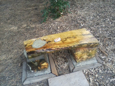 Smooth wooden bench outside with a painted rock sitting on it.