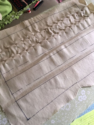 A beige rectangle of fabric with the edges marked with black sharpie and large seam allowances left outside the marks, with two pleats very neatly arranged and pinned across the edge of the pleat about every half inch, and two more pleats marked in black sharpie with dashed lines for the under fold and a solid line for the visible edge. The lower edge of the fabric is still creased in wavy lines from the first attempt at pleats.  The pile of sewing supplies on the couch, just visible, includes large silver shears, their black plastic sheath, a spool of red thread.