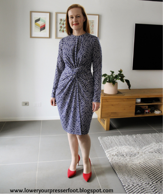 white woman posing in a blue twist front dress wearing red shoes standing in a lounge room