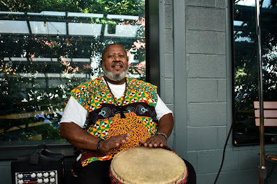 Photo of Barry Johnson. He is sitting down, wearing a brightly colored yellow and green vest. A drum rests between his legs and his tap lightly against the drum.