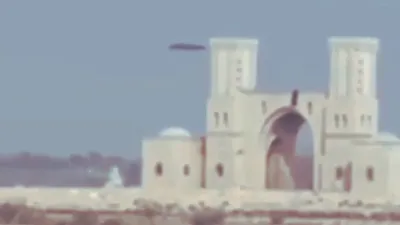 Silver disk UFO flying slowly behind Qatar Tribal Farm Gate with wind towers.