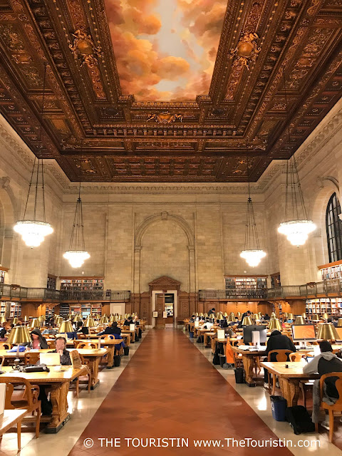 People sitting in about eight rows of wooden desks in a large reading room under an ornamented dark wooden coffered ceiling that has a pastel-coloured painting in its centre.