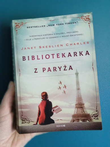 Na tle granatowej ściany dłoń trzyma książkę pod tytułem Bibliotekarka z Paryża autorstwa Janet Skeslien Charles.