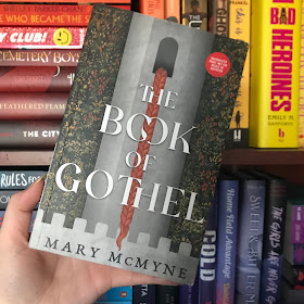 The Book of Gothel by Mary McMyne held up by a white hand in front of rainbow shelves.