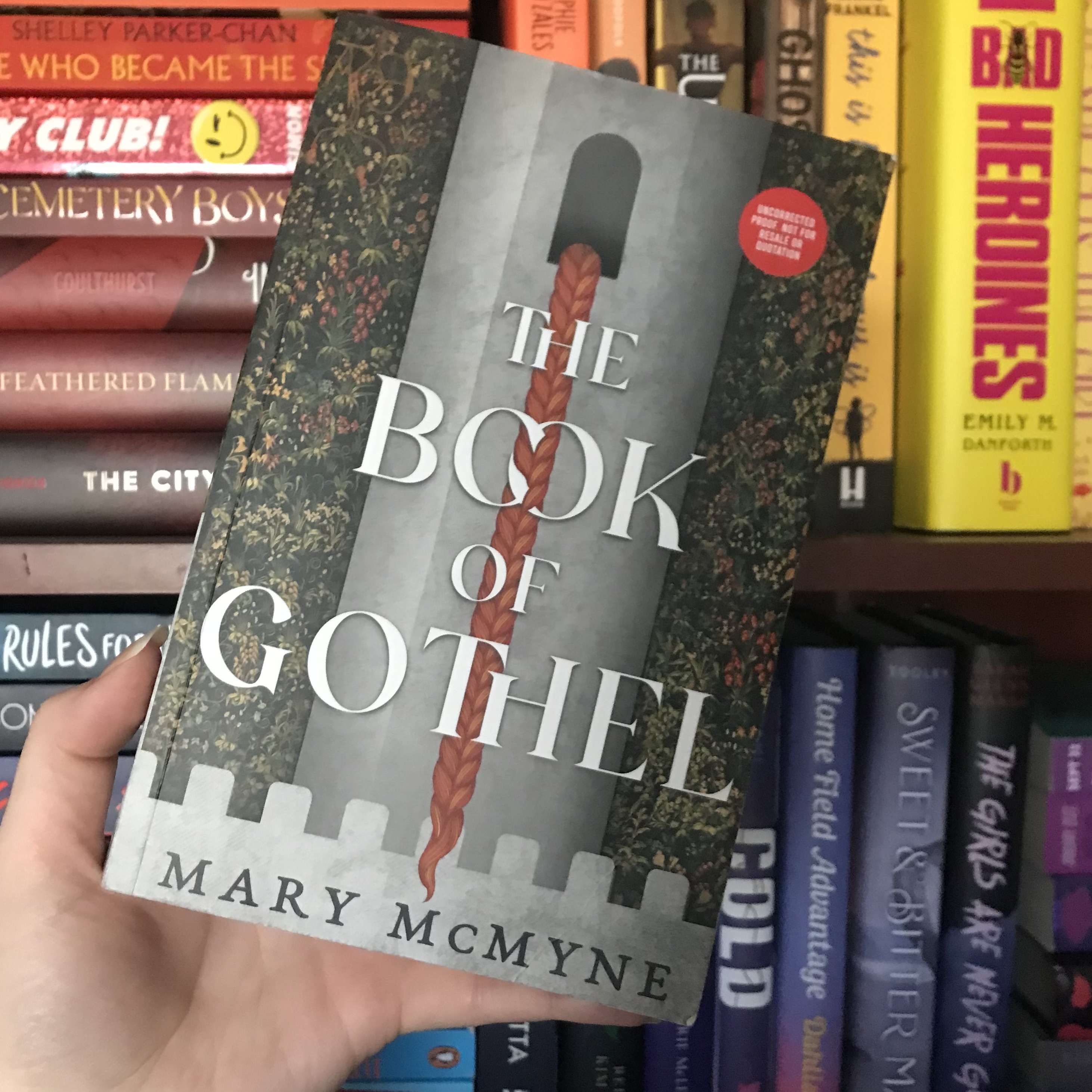 The Book of Gothel by Mary McMyne held up by a white hand in front of rainbow shelves.