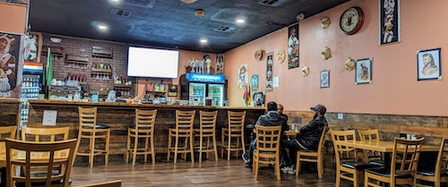 Interior scene showing bar and customers at one table with walls covered in photos and handicrafts