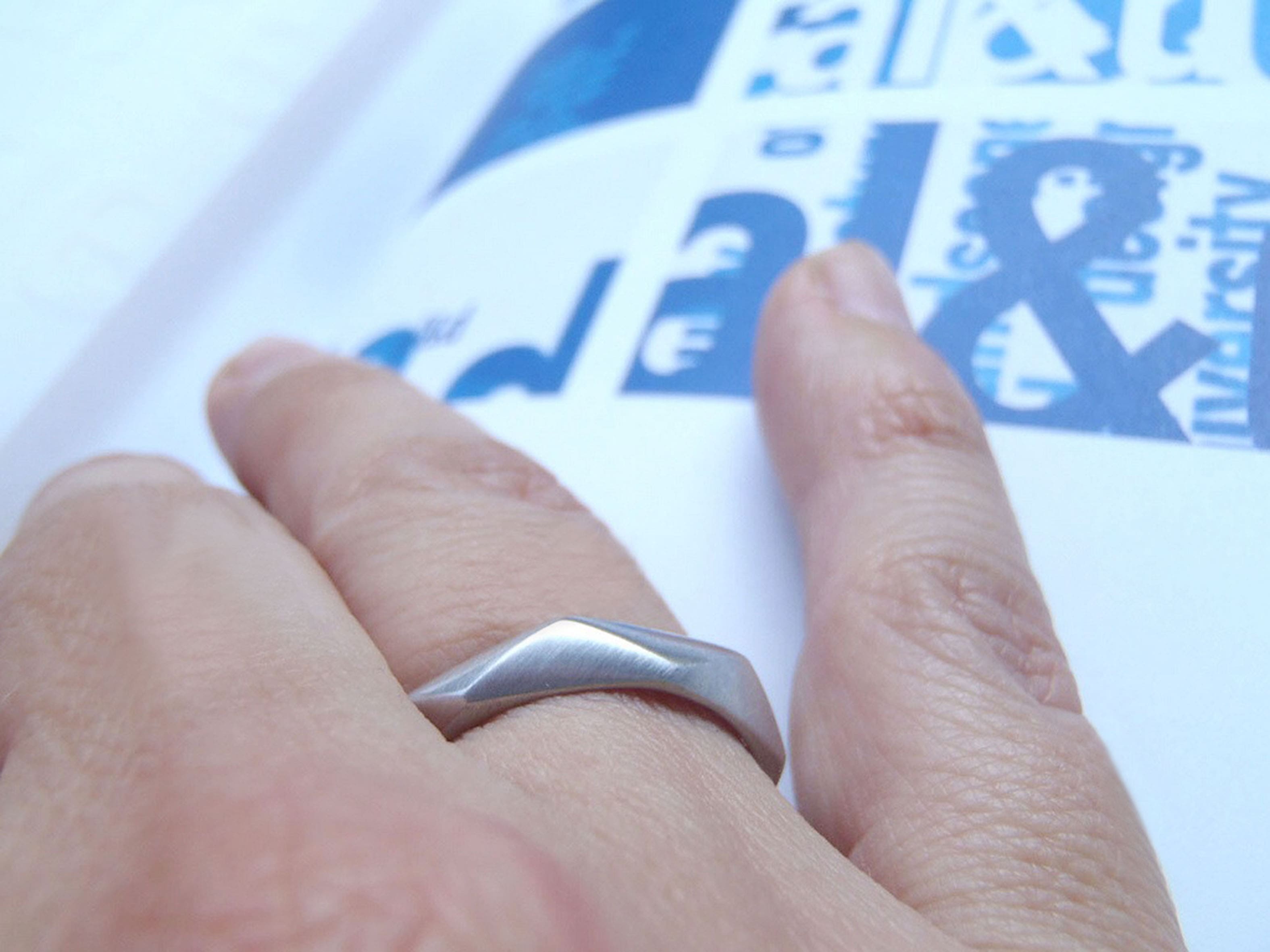 Una mano izquierda descansa sobre un la página de un libro en tono gris claro, lleva una  argolla de matrimonio de diseño geométrico minimalista hecha en oro blanco y con acabado rodizado mate
