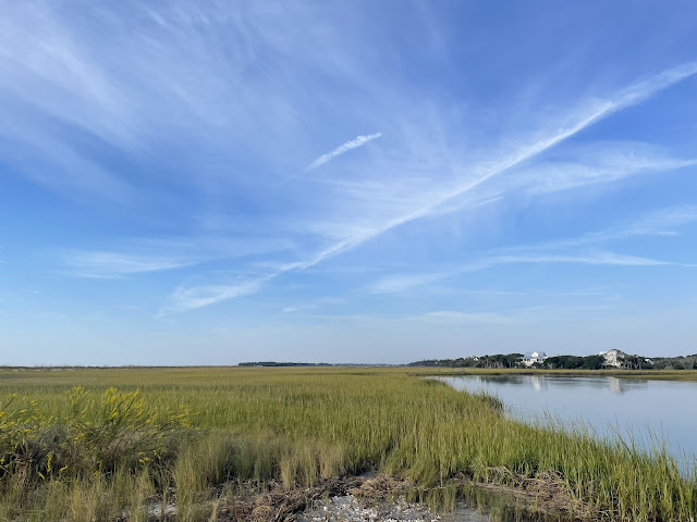 A view of the marsh that seperates the ocean from the land. The sky is blue with light white clouds streaking the sky. The march grass is green and wheat colored between knee and hip high. The tide is high so there is a lot of water in the channel seperating the land and the ocean.