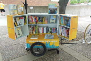 Book cabinet atop bicycle trailer, open to display cabinet shelves filled with books