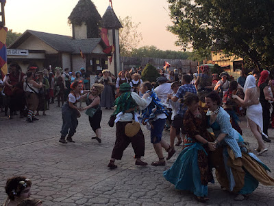 A crowd of dancers whirling in pairs over a cobbled courtyard in front of a plaster-and-timber gatehouse, in various combinations of Renaissance, pirate, and modern clothing, at sunset. The gold light of the sun just touches the buildings in the far background and the heads of some of the taller, further dancers; the rest are shadowed.