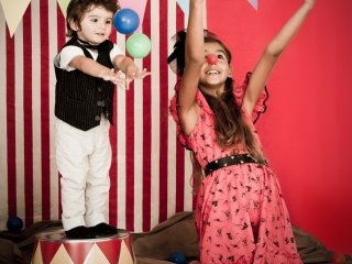 A boy juggles while a girl cheers with raised arms, wearing a red clown nose, as they perform a circus show.