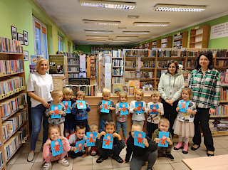 Zdjęcie grupowe przedszkolaków, pań bibliotekarek i pani przedszkolanki. Tło: lada biblioteczna i regały z książkami.