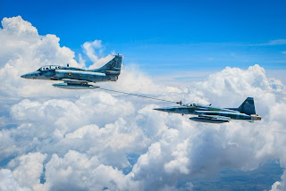 Descrição: Foto em dia de céu azul com nuvens: dois aviões caça em voo com proa à esquerda, a uma certa distância e em níveis diferentes. À frente e em nível mais alto, o A-4 Skyhawk da Marinha Brasileira, aeronave de ataque, bombardeiro e multifunção, com motor turbojato, com capacidade de abastecimento a mais de 22 000 pés com transferência de até 2.200 litros de combustível por minuto. O sistema de reabastecimento do caça A-4 da Marinha do Brasil estende a mangueira em direção ao F-5M da Força Aérea Brasileira,um caça leve, com motores turbojato, bimotor monoplano, projetado na aplicação de aviônicos de última geração, nos sistemas de navegação, armamentos e auto-defesa. O contato é feito em pleno ar, no final da mangueira, situa-se o drogue, acoplador com forma afunilada, que é estabilizado por uma cesta. A aeronave recebedora possui um probe (na parte externa, preso a fuselagem, à direita próximo ao cockpit envidraçado) que acopla o drogue e permite a transferência de combustível em voo.