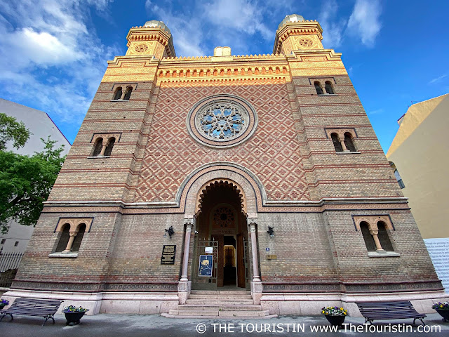 The facade of a red and cream-coloured synagogue decorated in Moorish style with a gigantic rose window over the entrance under a blue sky.