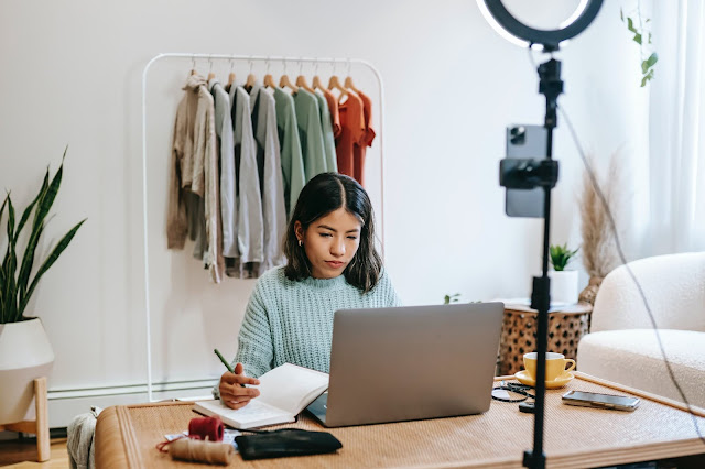 A brunette lady working on her laptop. A rack of clothes stand behind her.