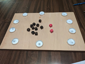 Ten used tea light candles in a circle on a wooden board, with several black dice and two red dice in the centre of the circle.