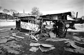 A black and white photograph of the trashed shanties. A banner reading "Racists did this" has been hung on the wreckage.