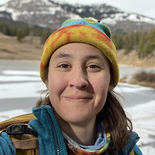 A picture of geologist Mara Reed, she is out in nature near some mountains wearing a yellow and blue beanie.